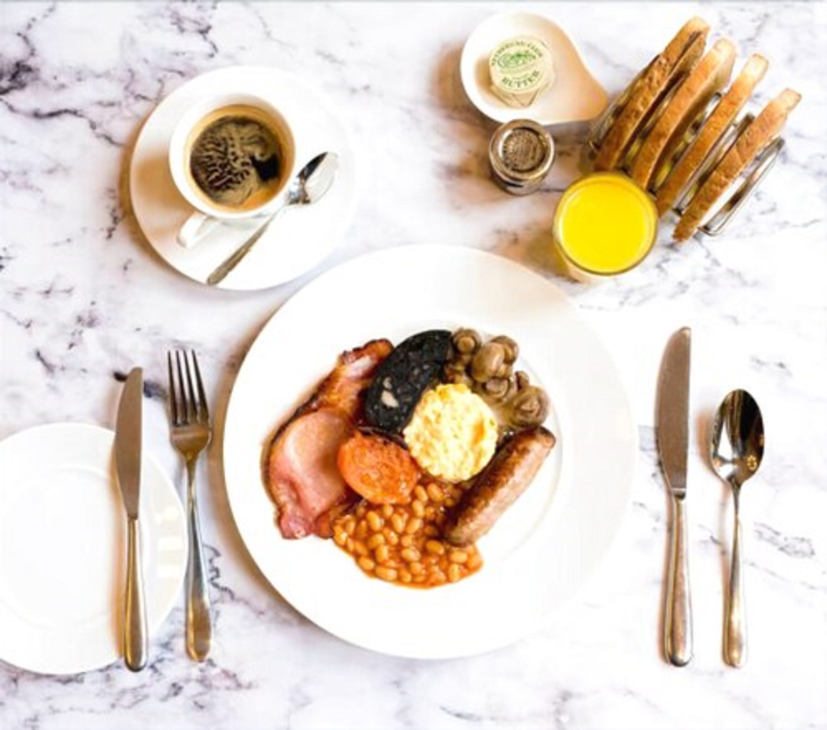 A breakfast setting with a plate of eggs, bacon, beans, and mushrooms, accompanied by toast, butter, and a cup of coffee.