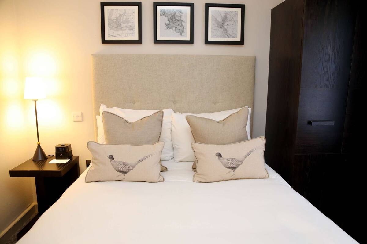 A neatly made bed with two pillows featuring bird designs, a bedside lamp, and a nightstand with a clock. Three framed artworks are displayed on the wall above the bed.