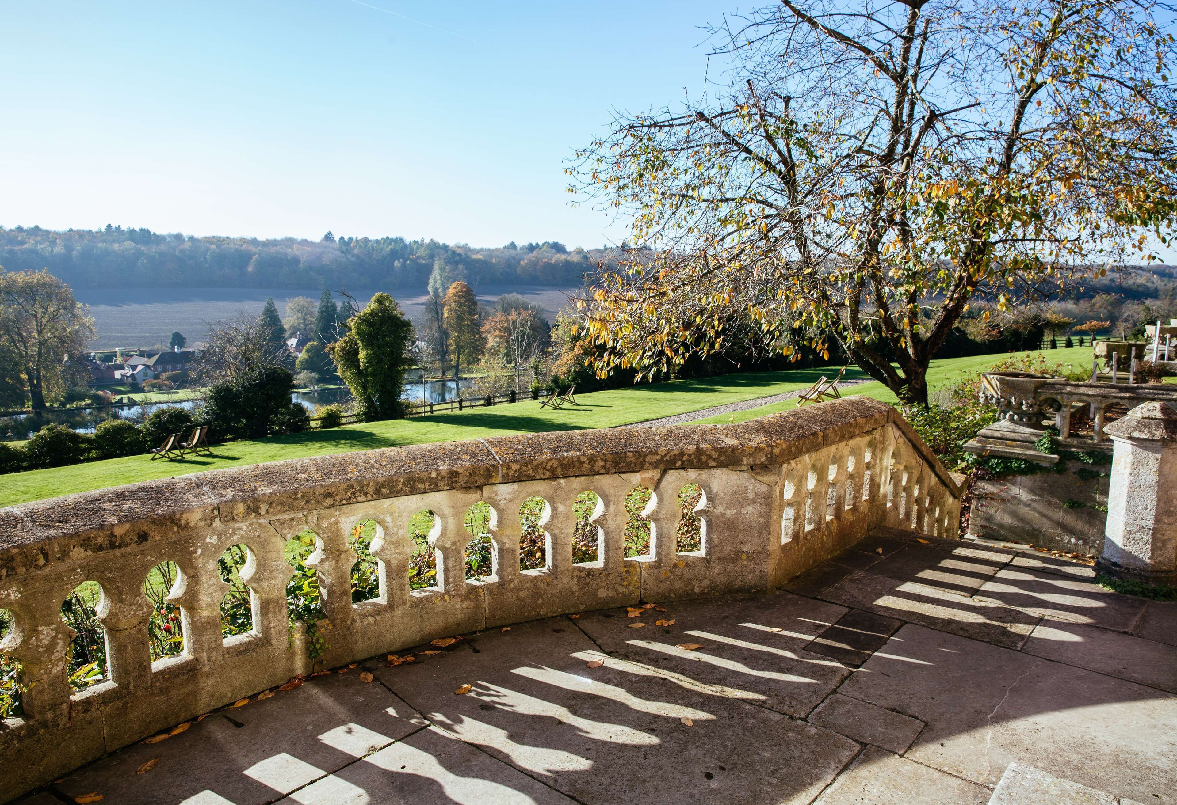 A stone balustrade with arched openings overlooking a park with a lake, trees, and distant buildings.