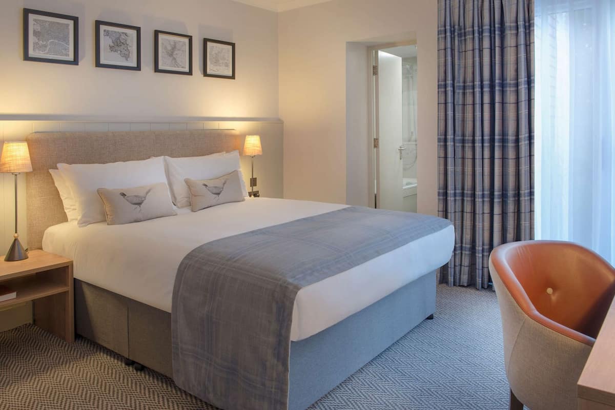 A hotel room with a bed, two bedside lamps, a chair, a nightstand, and a door leading to a bathroom.
