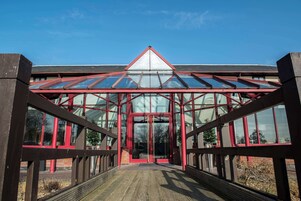 Entrada a un invernadero con techo de vidrio y estructura roja, que da paso a un edificio de ladrillo.