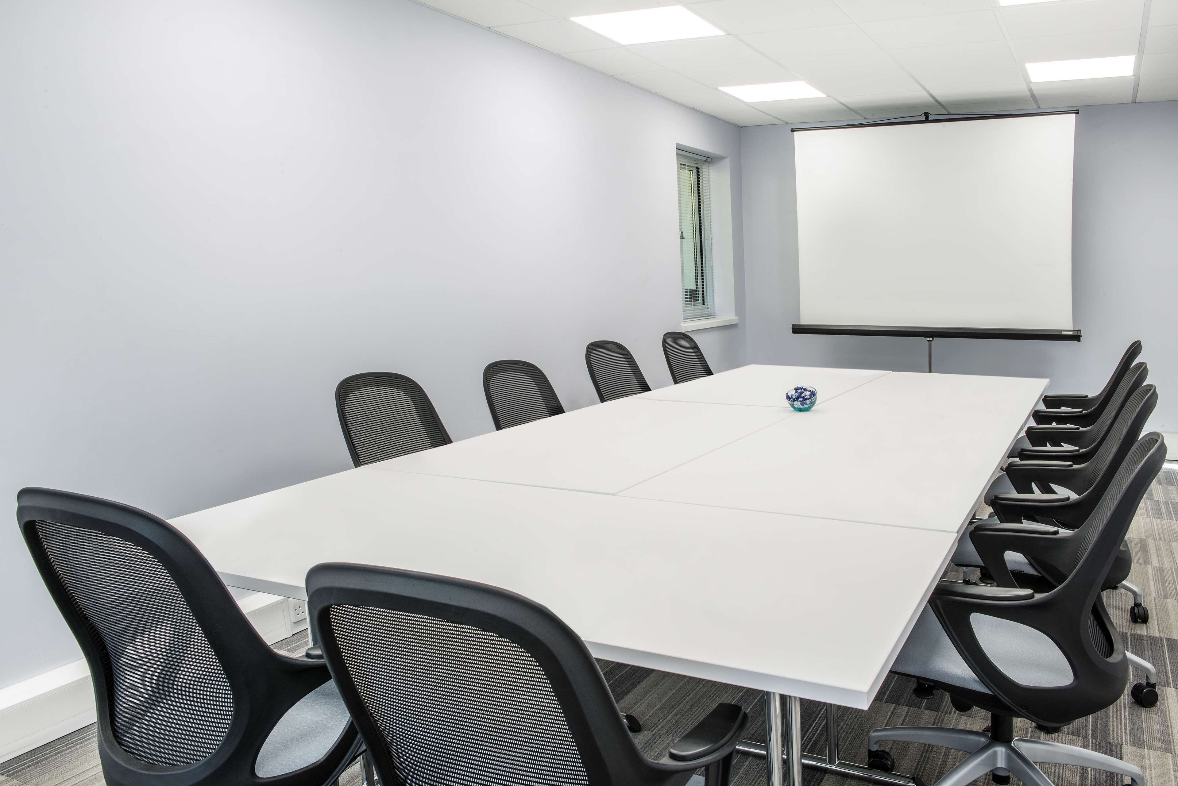 Un salón de conferencias con una mesa blanca, sillas de malla negra, una pantalla de proyector y una ventana con persianas.