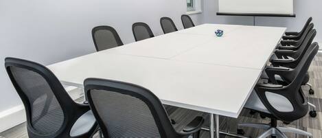 Un salón de conferencias con una mesa blanca, sillas de malla negra, una pantalla de proyector y una ventana con persianas.