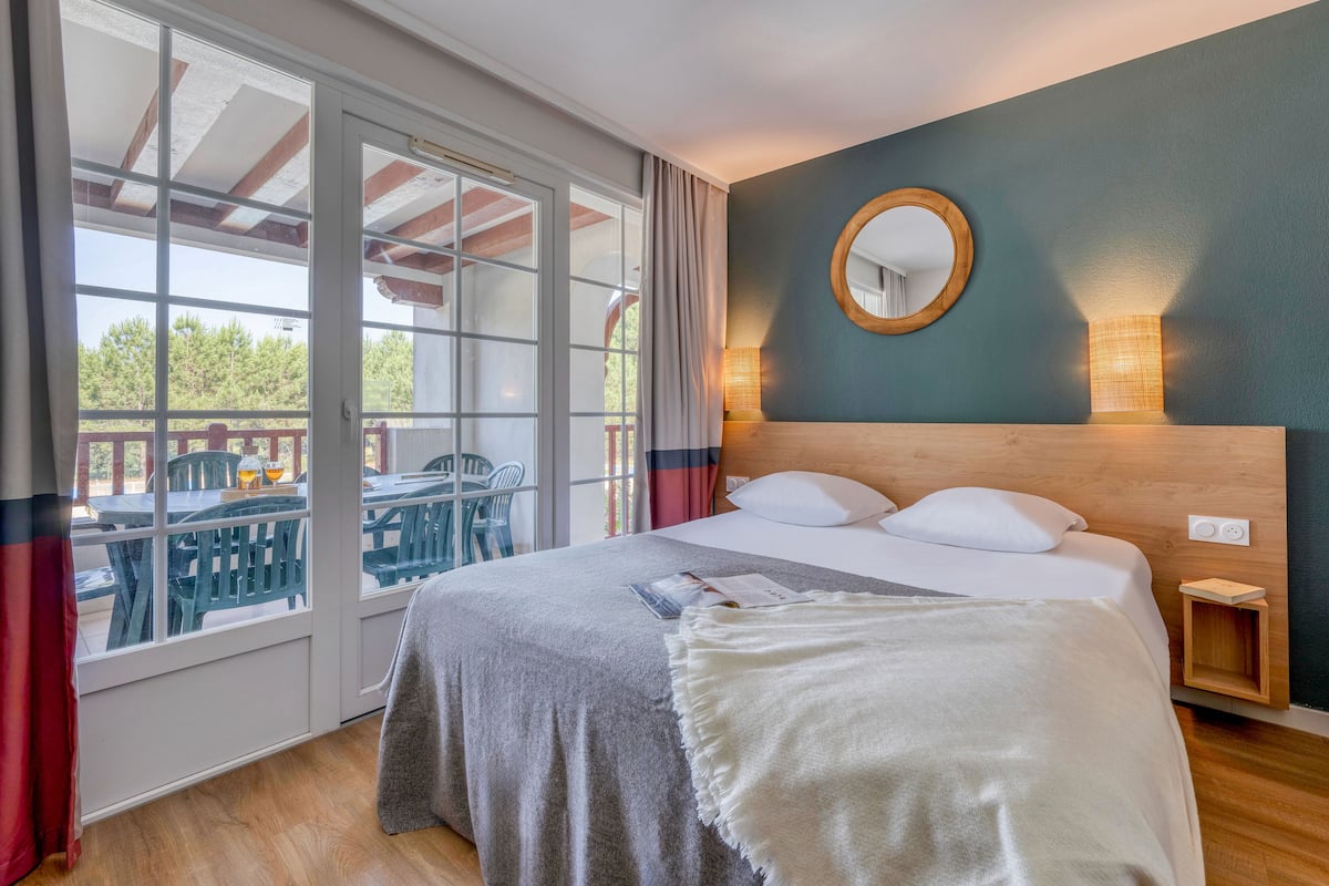 A bedroom with a large bed, a wooden headboard, a nightstand, a mirror, and a view of an outdoor deck with chairs and a table.