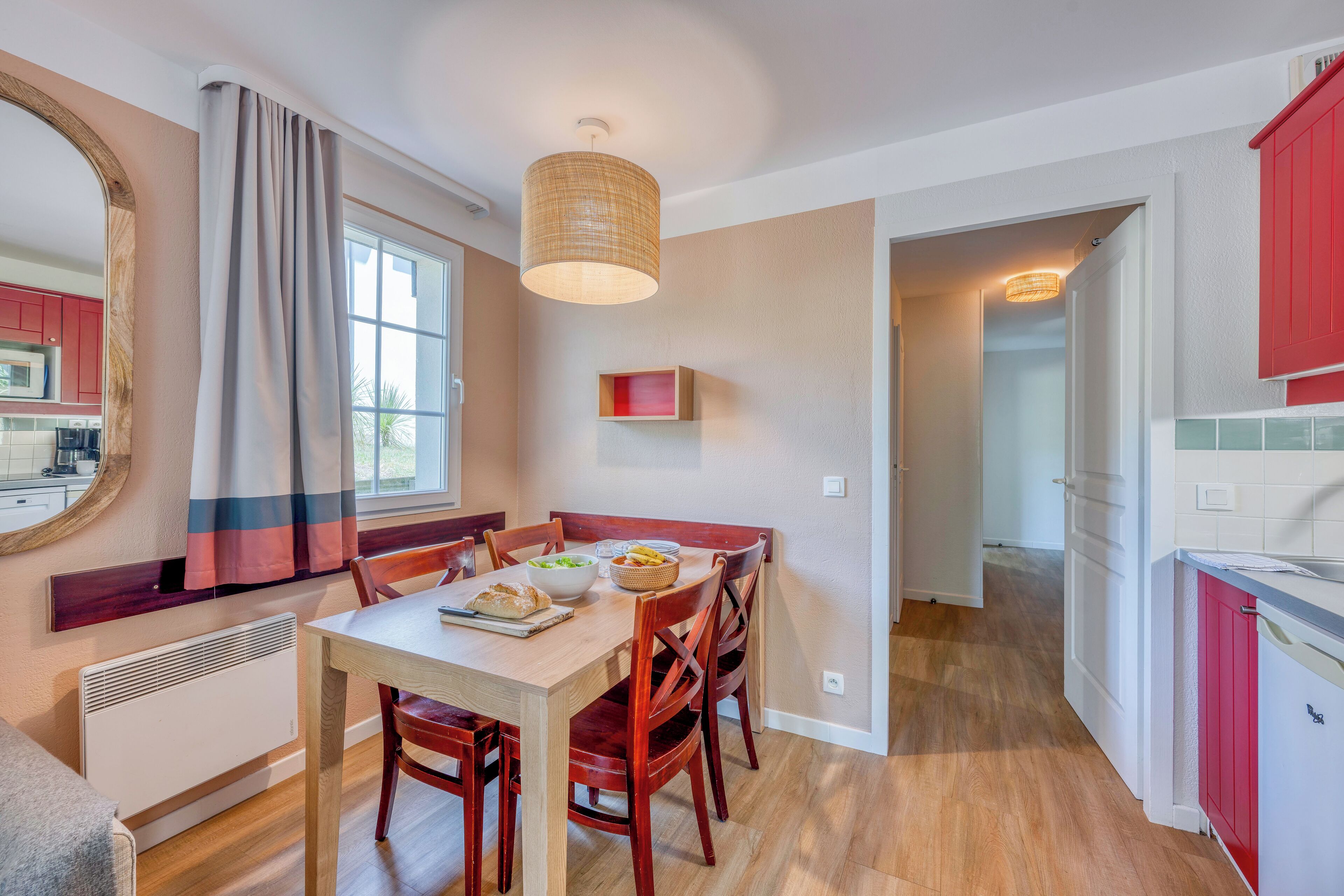Un espace repas comprenant une table, des chaises et un luminaire suspendu. On y trouve également une cuisine avec des placards rouges et une fenêtre.