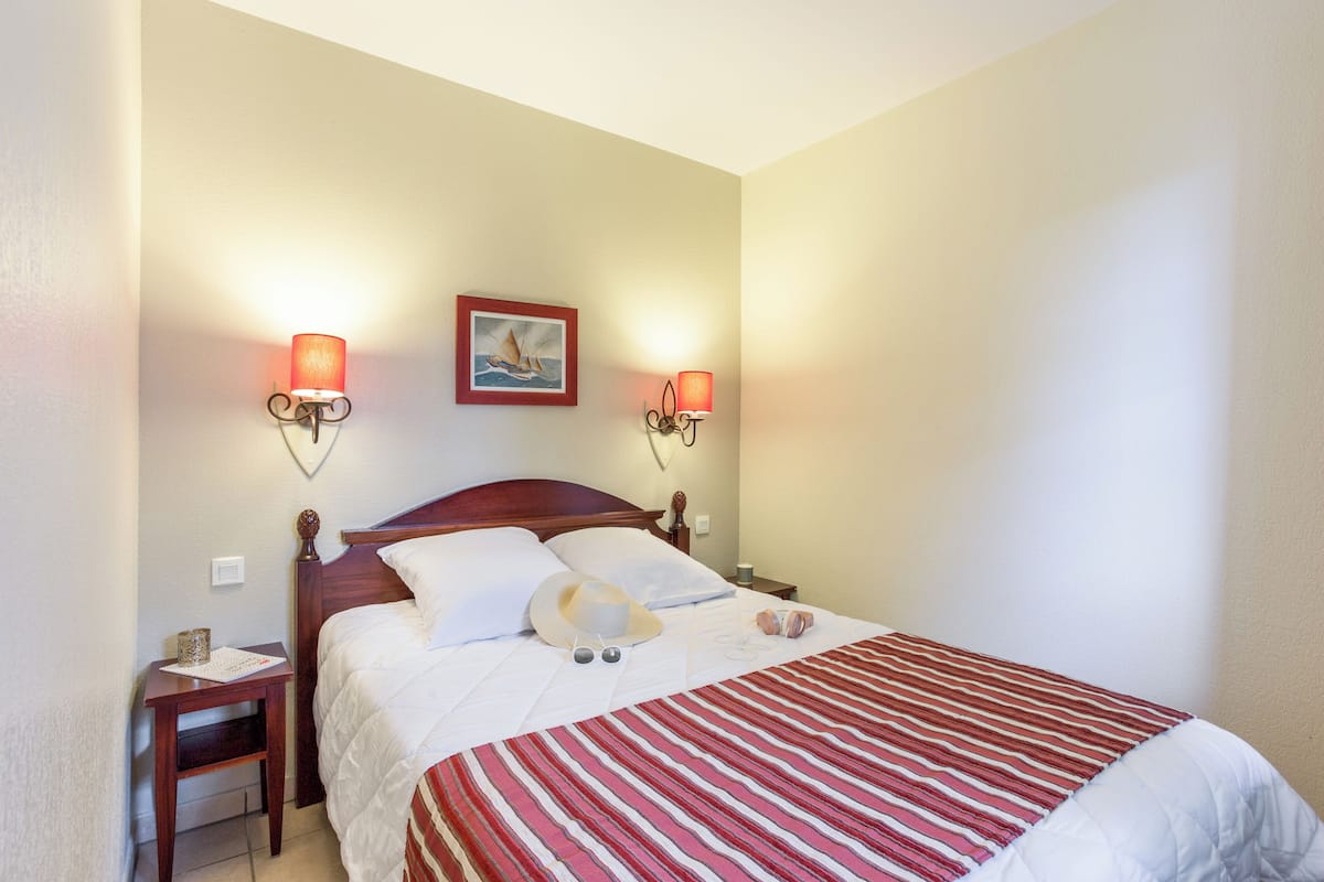 A neatly made bed with a striped bedspread, two wall-mounted reading lamps, a framed picture on the wall, and a small bedside table with a lamp and a book.