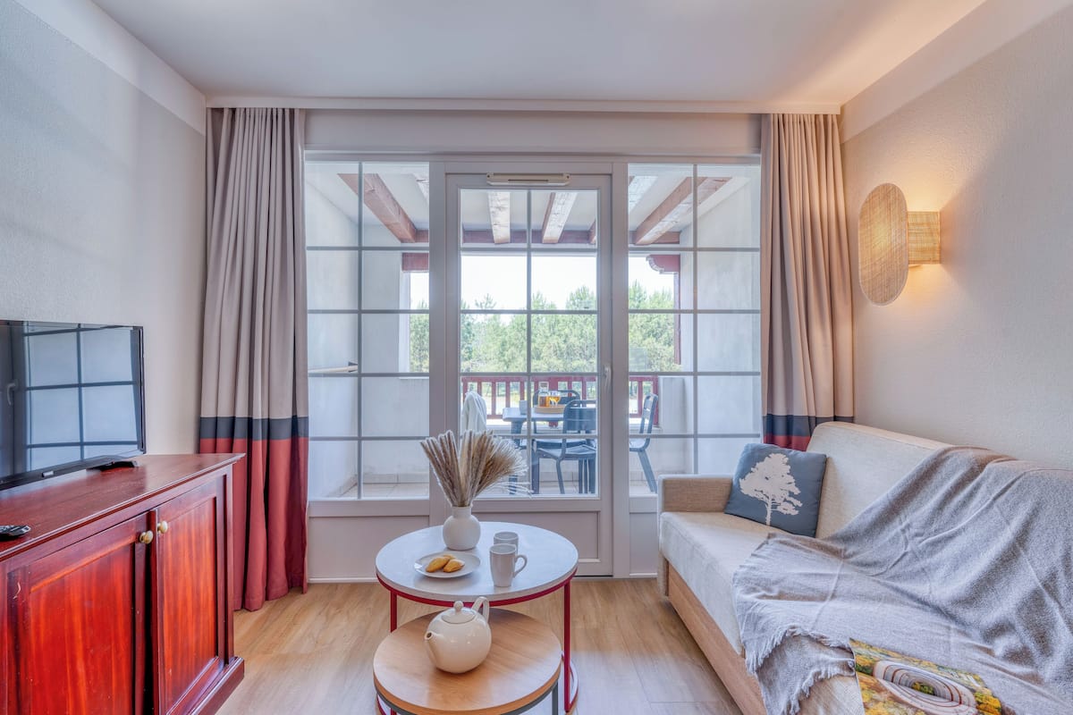 A cozy living room with a sofa, a small table with a vase and cups, a TV on a cabinet, and a large window with curtains.