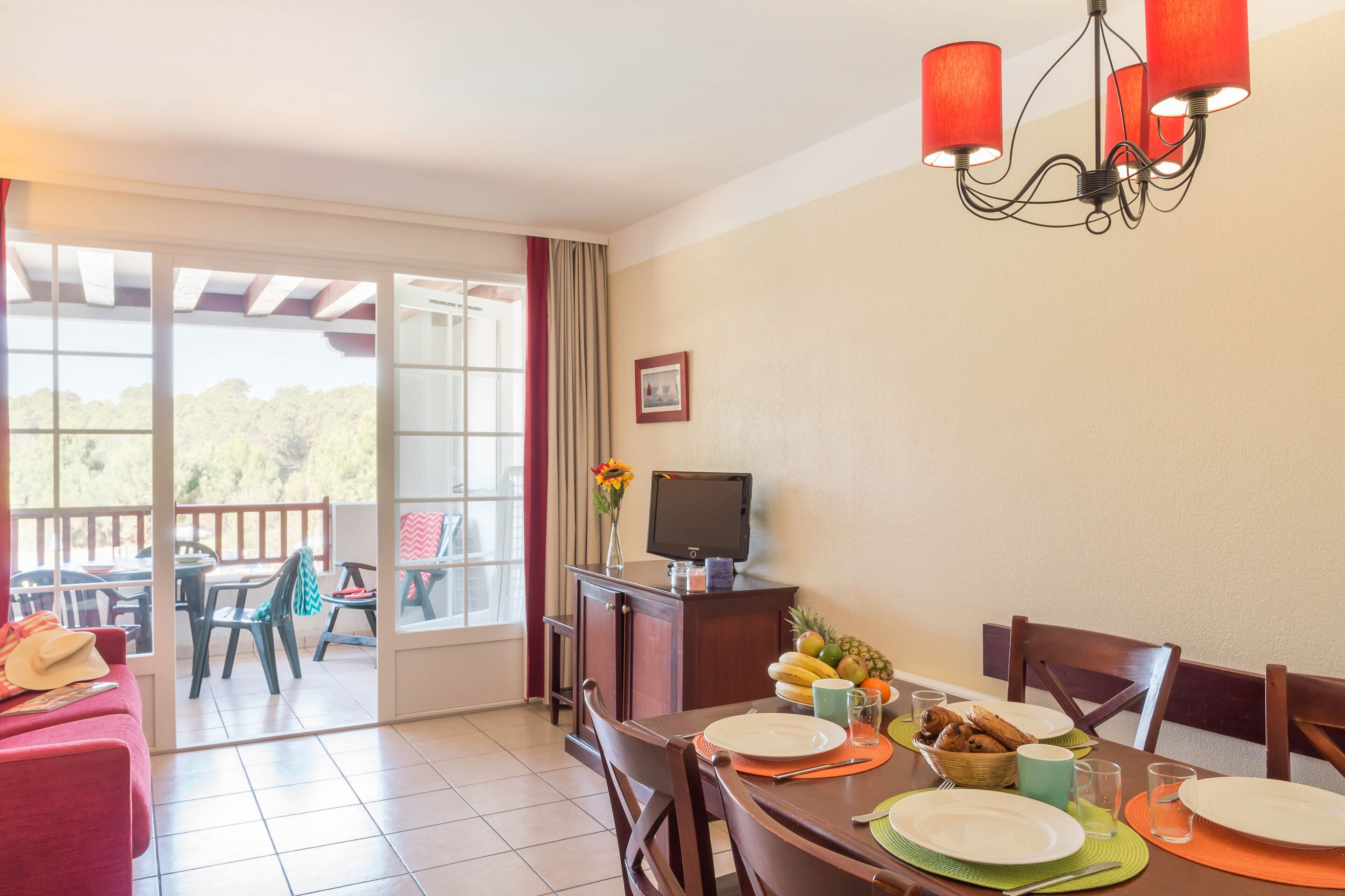 Un comedor con una mesa dispuesta para dos, un televisor sobre un mueble y vistas a un patio exterior a través de un ventanal.