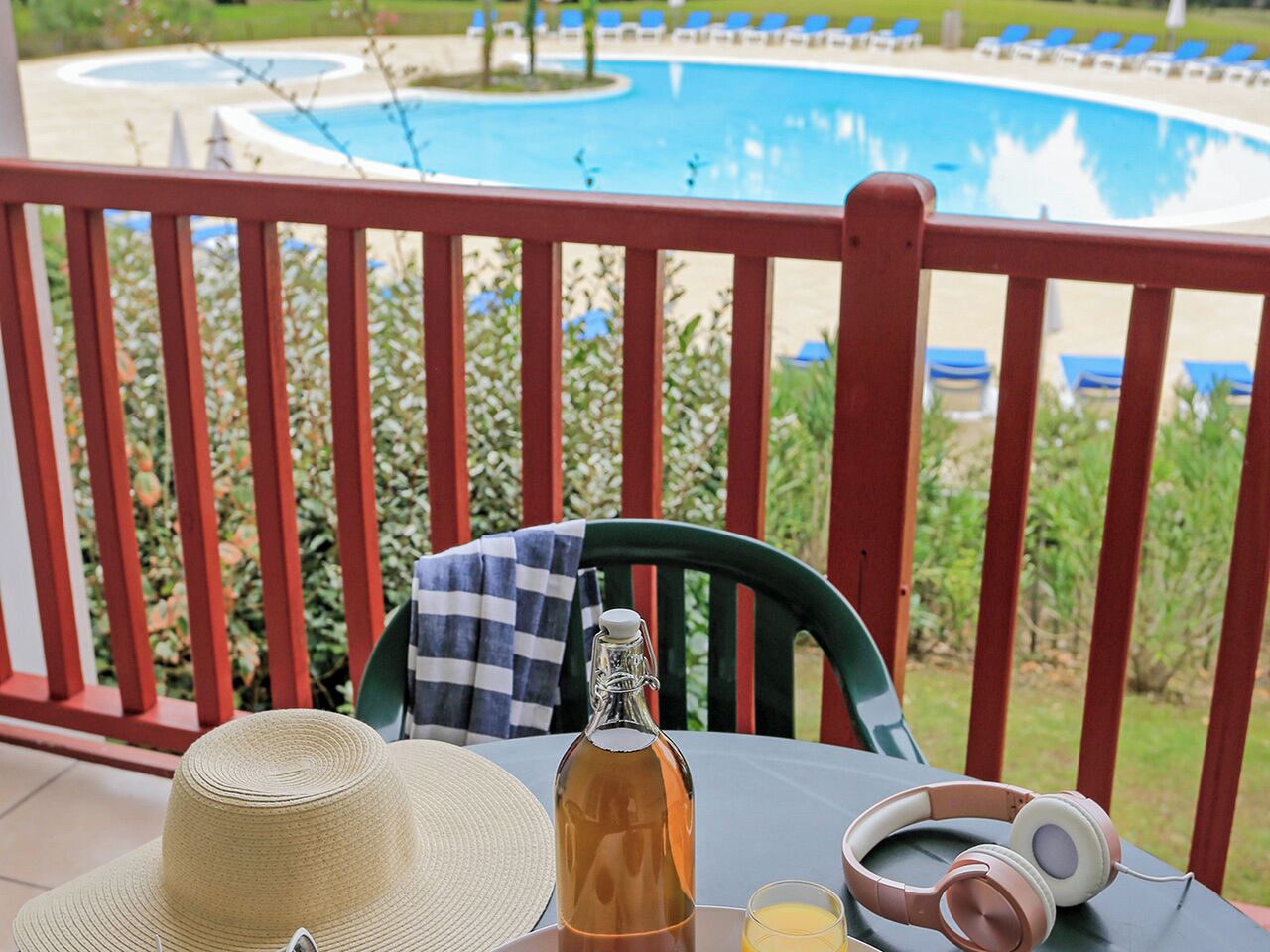 A table with a bottle, a straw hat, and headphones on a balcony overlooking a pool.
