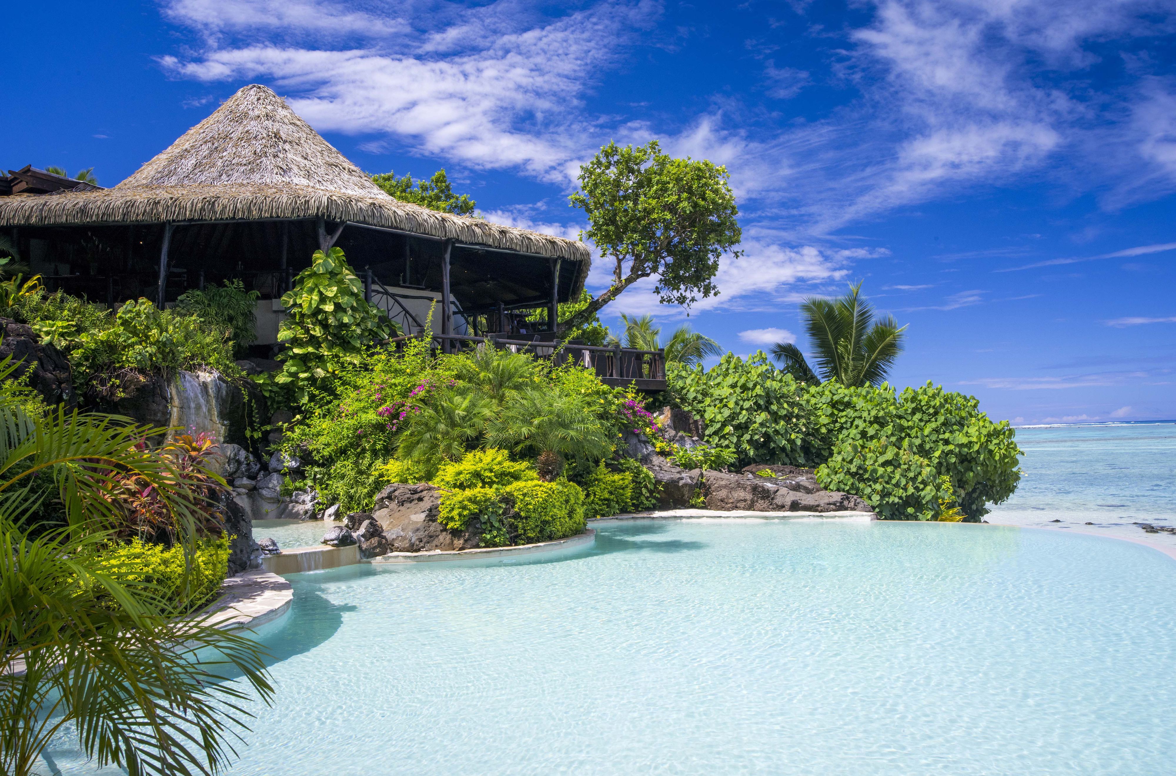 Un resort con una construcción de techo de paja, piscina y abundante vegetación.