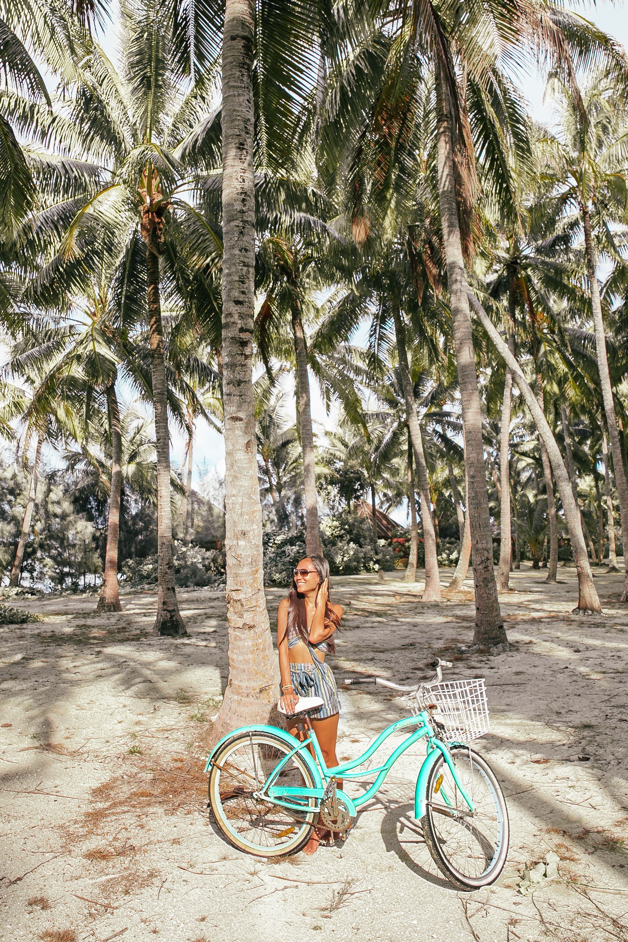A turquoise bicycle parked under a palm tree in a sandy area with other palm trees in the background.