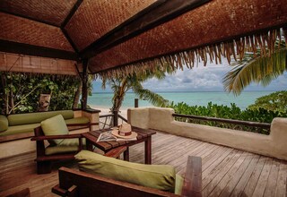 A wooden deck with a thatched roof, green cushioned seating, and a view of the ocean.