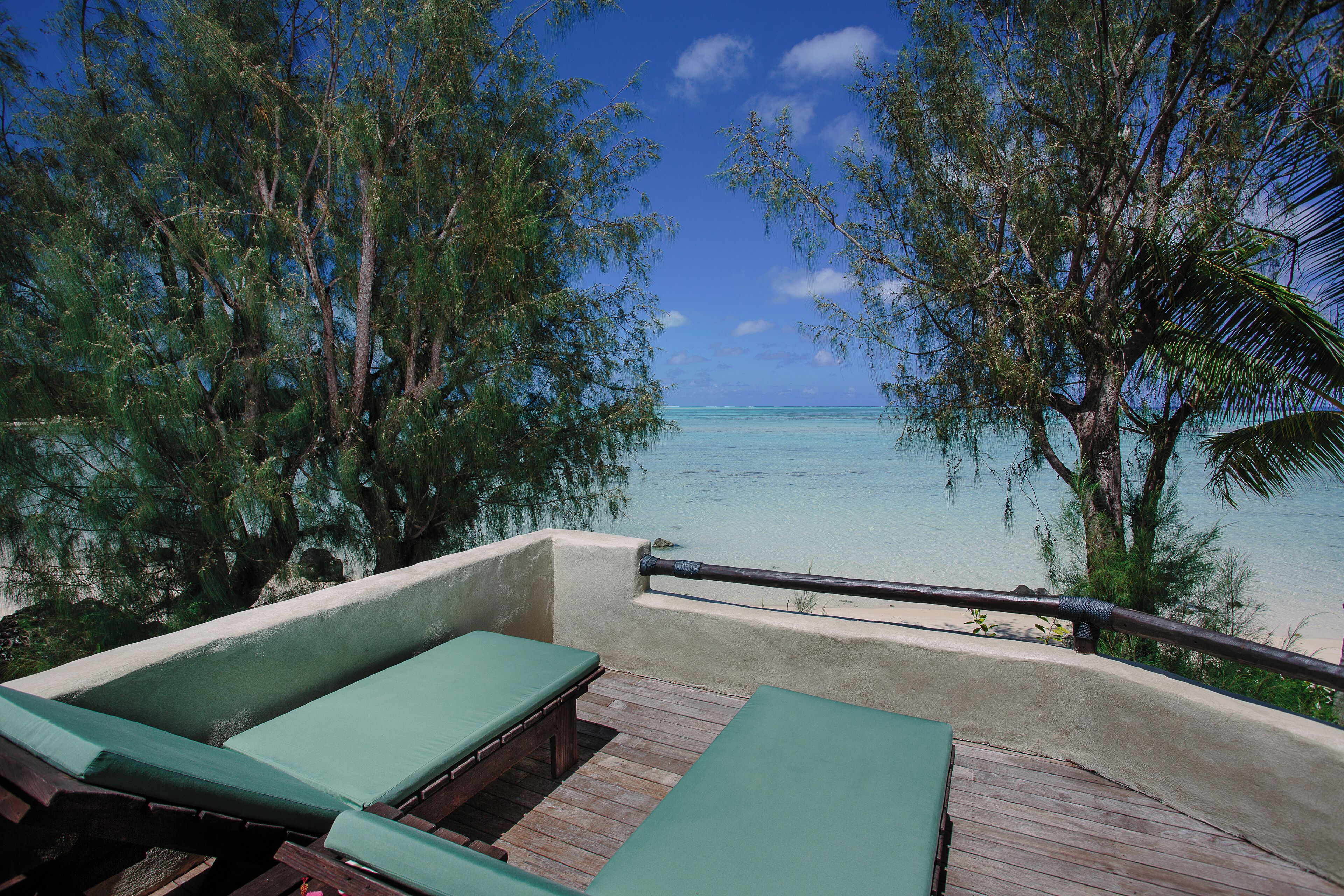 A wooden deck with green lounge chairs overlooking a clear blue sea, surrounded by trees.