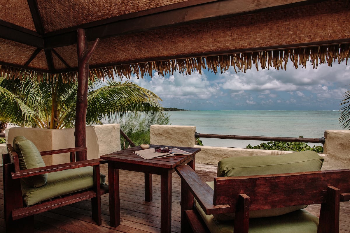 A wooden deck with a thatched roof, overlooking a turquoise sea and palm trees.