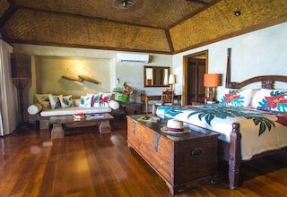 A room with a wooden floor, a bed with a floral bedspread, a wooden dresser, a sofa with colorful cushions, and a ceiling fan.