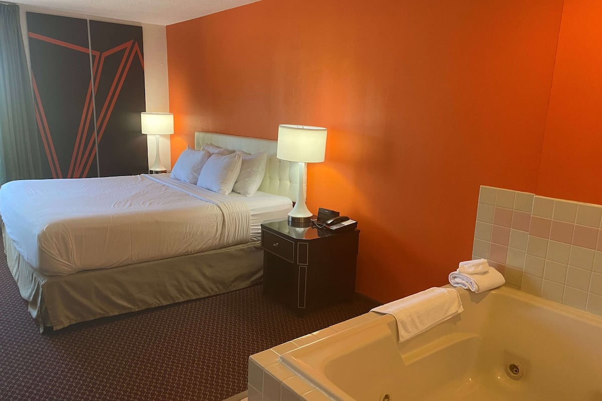 Een hotelkamer met een groot bed, twee nachtkastjes met lampen, een klein tafeltje met een telefoon en afstandsbediening, en een badkuip met een handdoek.