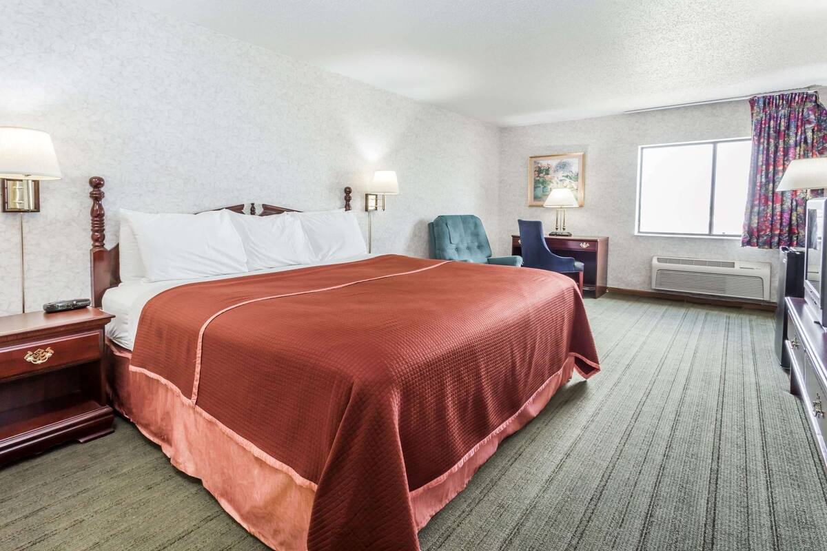 Een hotelkamer met een groot bed, twee nachtkastjes, een stoel, een bureau, een lamp, een schilderij en een raam met gordijnen.