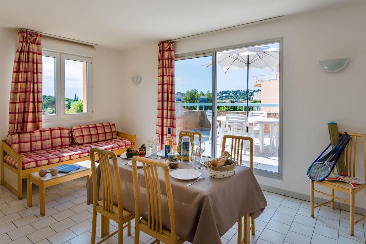 A dining area with a table set for two, a sofa, and a tennis racket on a chair.