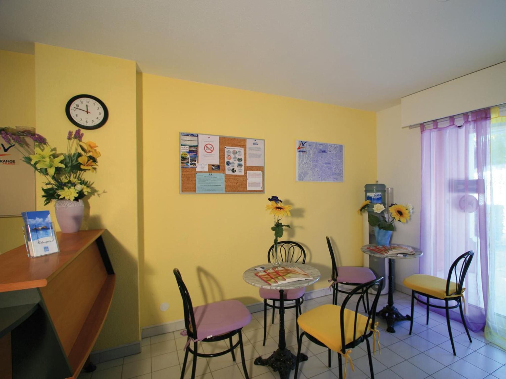 A dining area with a corkboard displaying notices, a clock, and a flower arrangement.