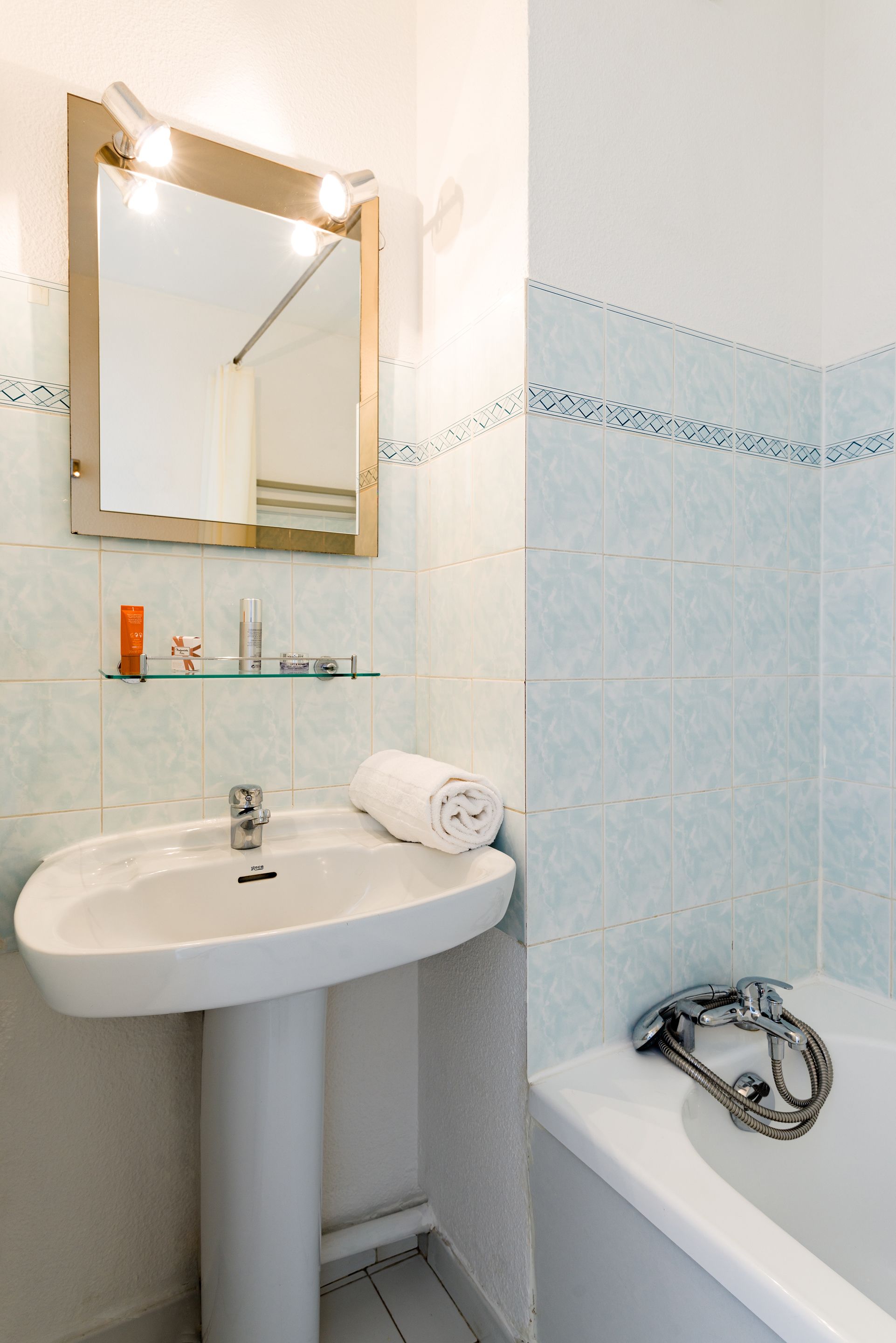 Ein Badezimmer mit einem weißen Waschbecken, einem beleuchteten Spiegel, einer Dusche und hellblauen, gemusterten Fliesen.