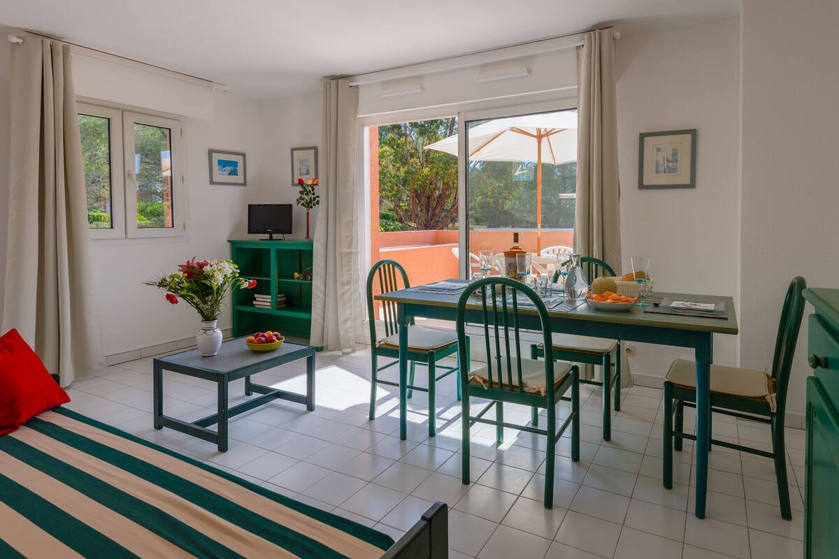 Een kamer met een bed, een eettafel met stoelen, een bijzettafeltje met een bloemenvaas, en een televisie op een dressoir.