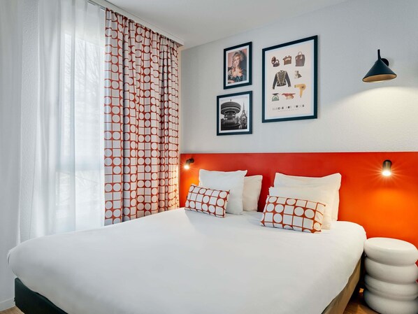 A neatly made bed with white bedding and patterned pillows, a red accent wall, and wall-mounted artwork including a cityscape and a fashion label.
