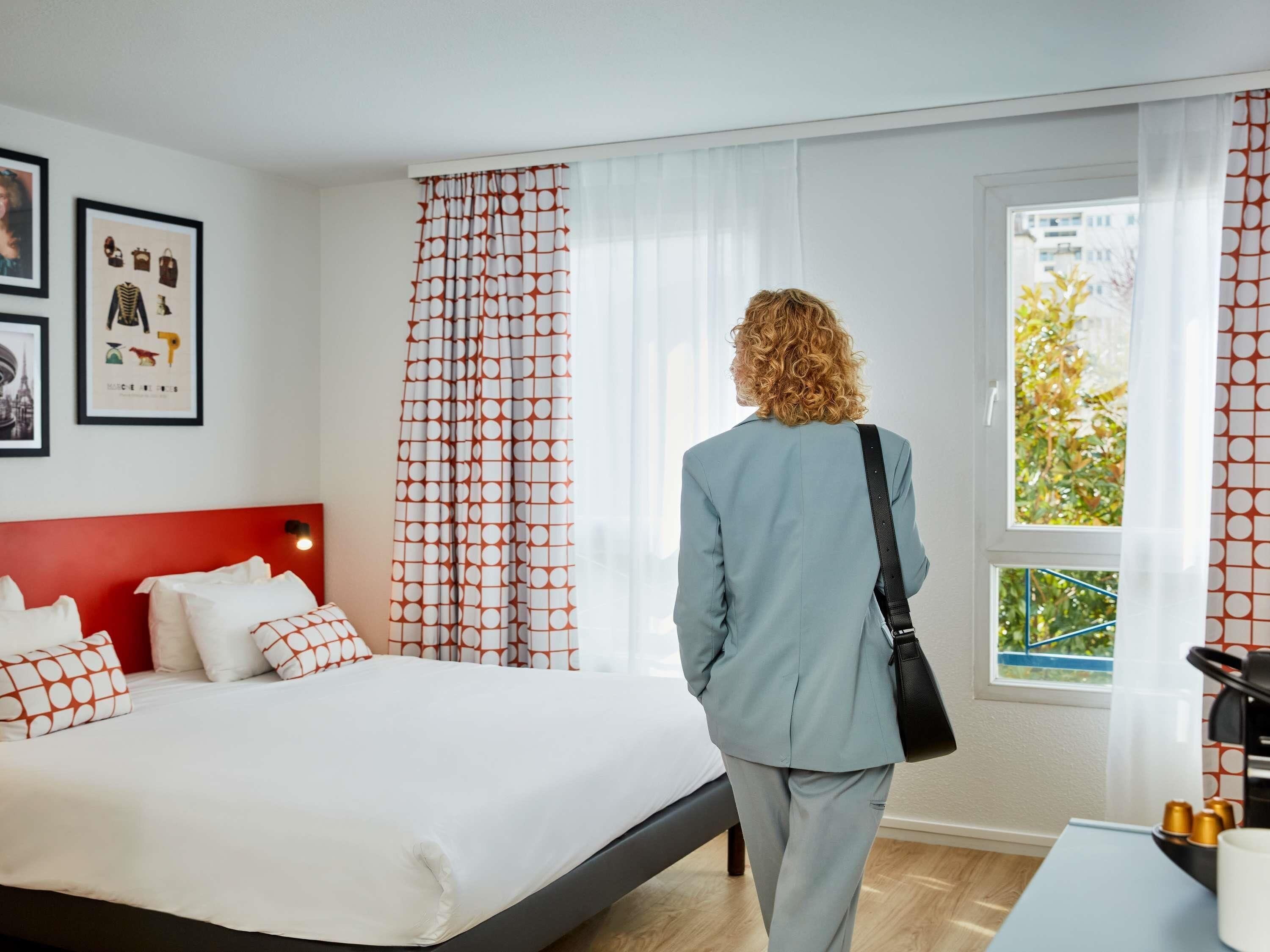 A hotel room with a bed, a window with curtains, a bedside table, and a wall with framed pictures.