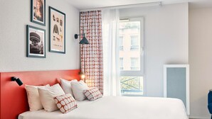 A modern bedroom with a red headboard, a large bed with patterned pillows, a bedside table with a book, and a window with a view of buildings. - Aparthotel Adagio Access Paris Vanves Porte de Versailles (Vanves)
