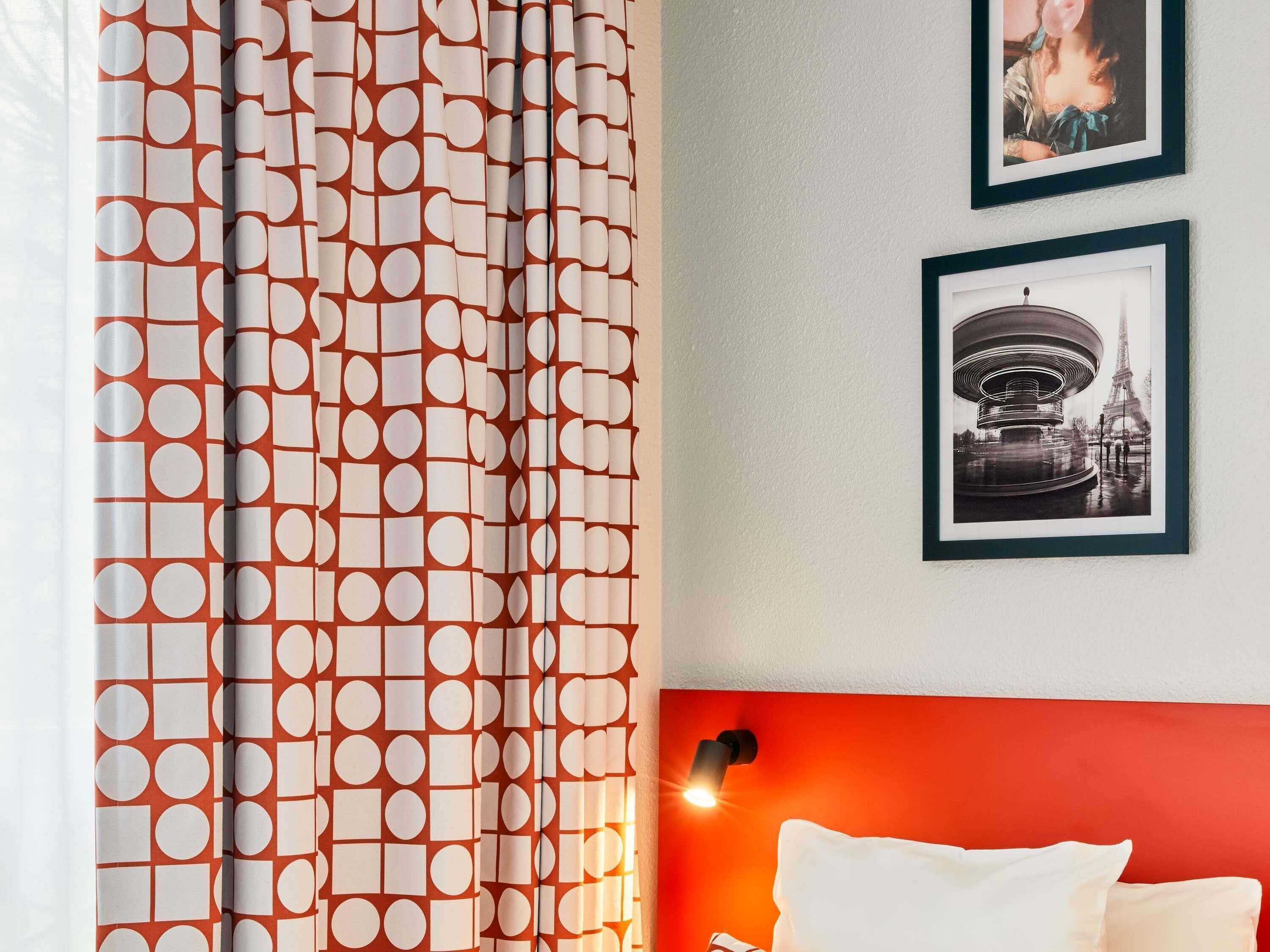 A hotel room with a patterned curtain, a bed with white pillows, and two framed pictures on the wall.