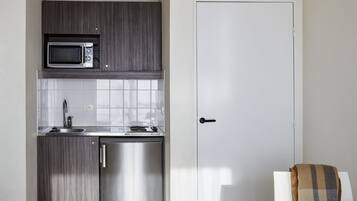 Een kleine keuken met een magnetron, spoelbak en vaatwasser, een witte deur, en een houten tafel met een stoel.