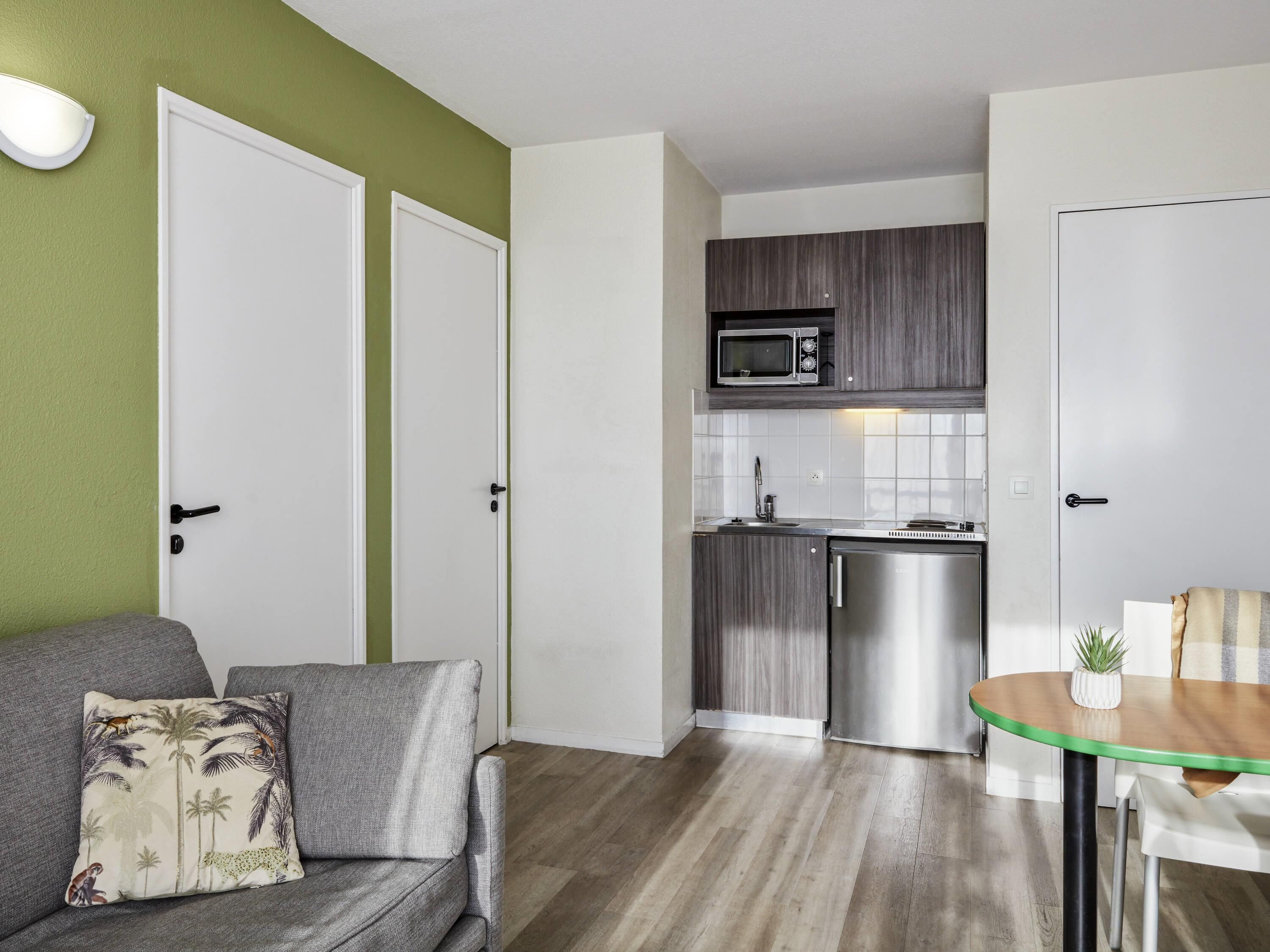 A compact living space with a kitchenette, a grey sofa with a tropical print pillow, and a small round table with a plant.
