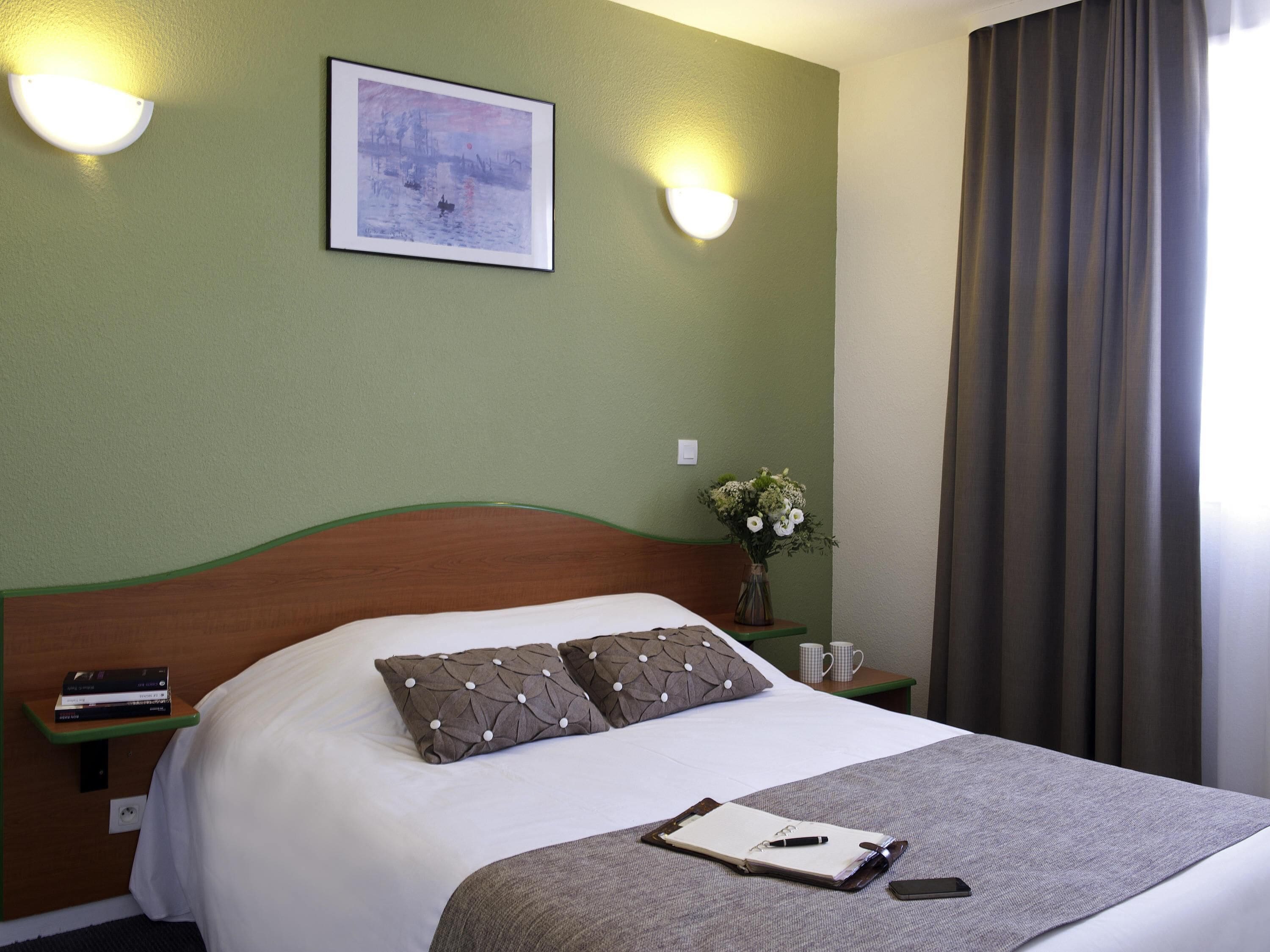 Een net opgemaakt bed met kussens, een nachtkastje met een boek en een bloemenvaas, en een schilderij aan de muur.
