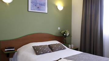 Een net opgemaakt bed met kussens, een nachtkastje met een boek en een bloemenvaas, en een schilderij aan de muur.