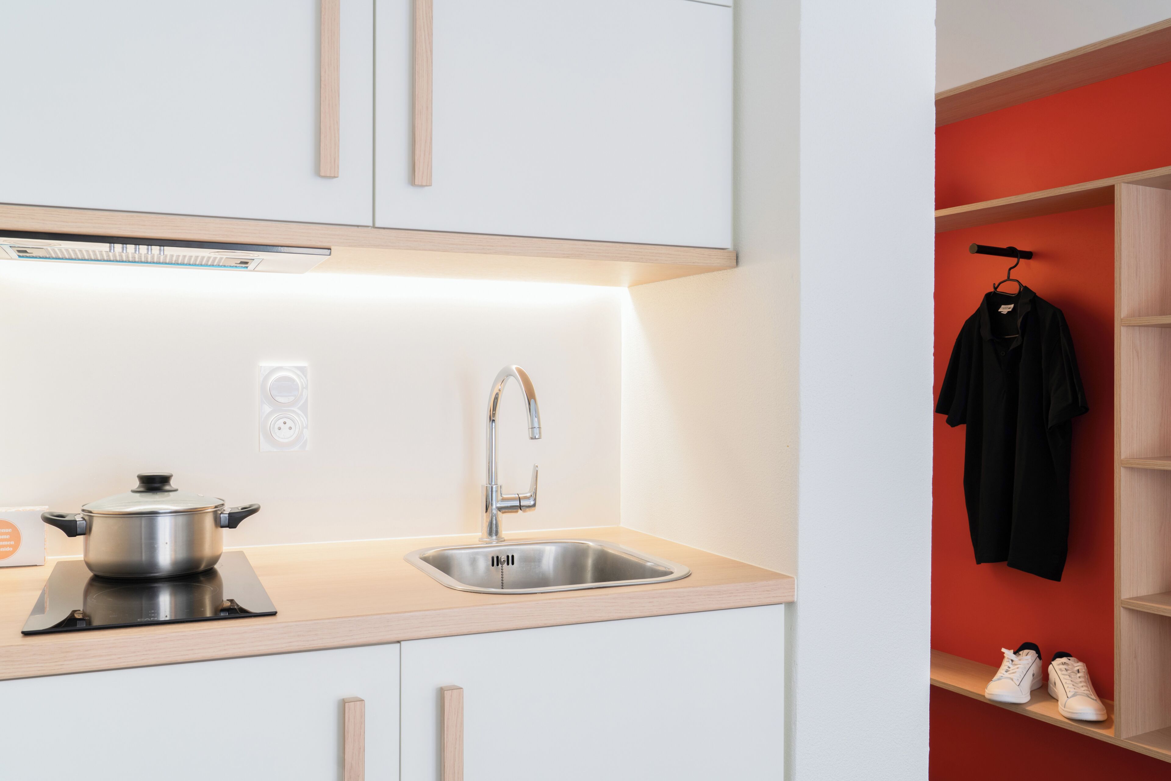 Una cocina moderna con un lavamanos de acero inoxidable, una olla sobre una cocina por inducción, y una pared roja con una chaqueta negra y zapatillas blancas en un estante.