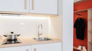 A modern kitchen with a stainless steel sink, a pot on an induction cooktop, and a red wall with a black jacket and white sneakers on a shelf.