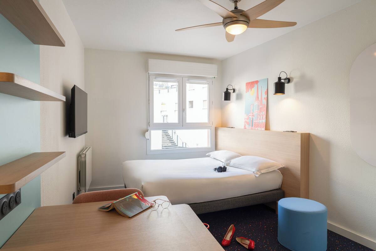 Ein Hotelzimmer mit einem Bett, einem Schreibtisch, einem Stuhl, einem Deckenventilator, einem Fenster und einem an der Wand montierten Fernseher.