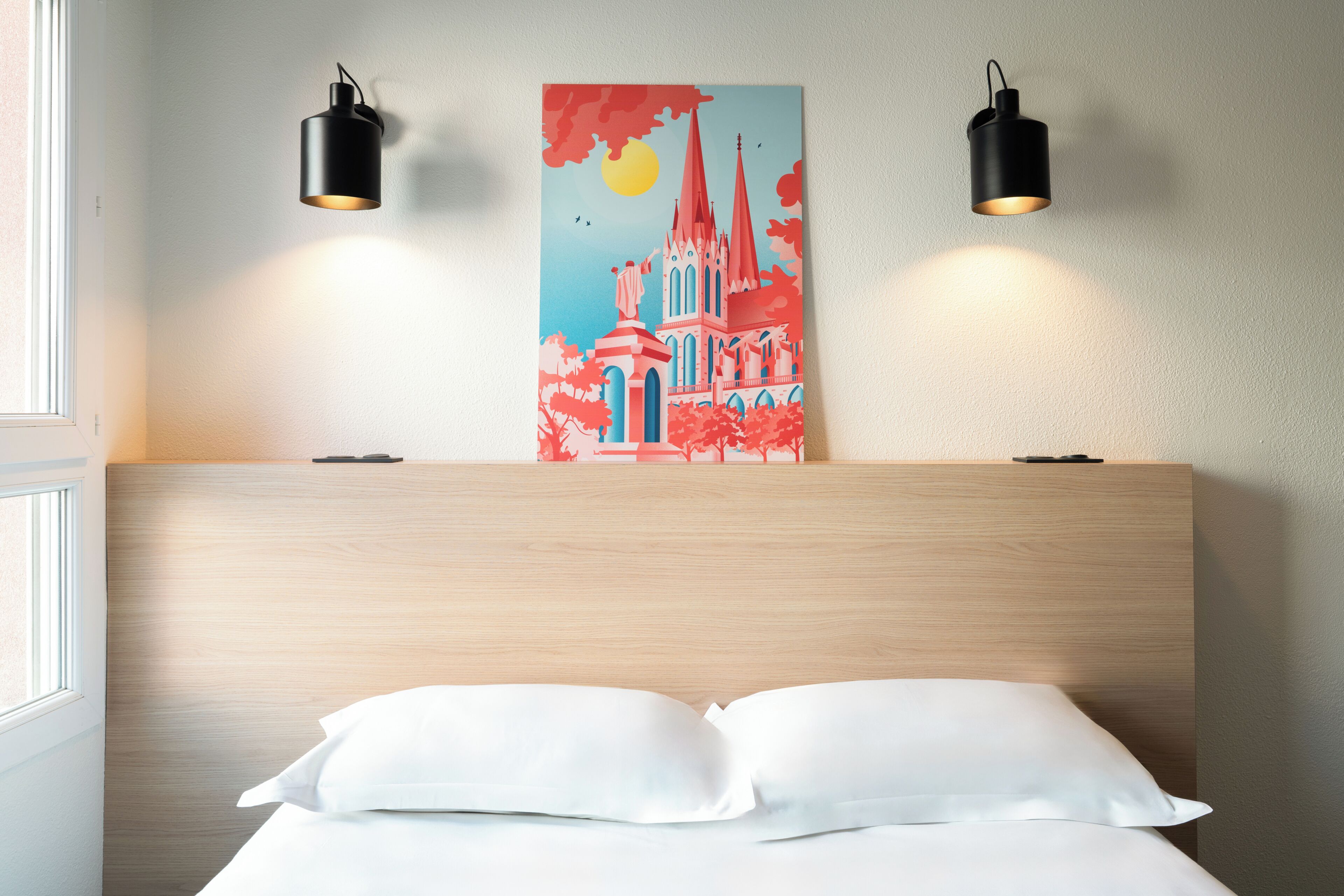 Ett hotellrum med en sänggavel i trä, två svarta vägglampor, en tavla som föreställer en byggnad och en vit säng med kuddar.