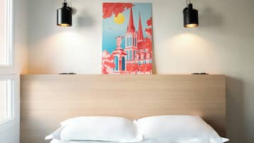 Ett hotellrum med en sänggavel i trä, två svarta vägglampor, en tavla som föreställer en byggnad och en vit säng med kuddar.