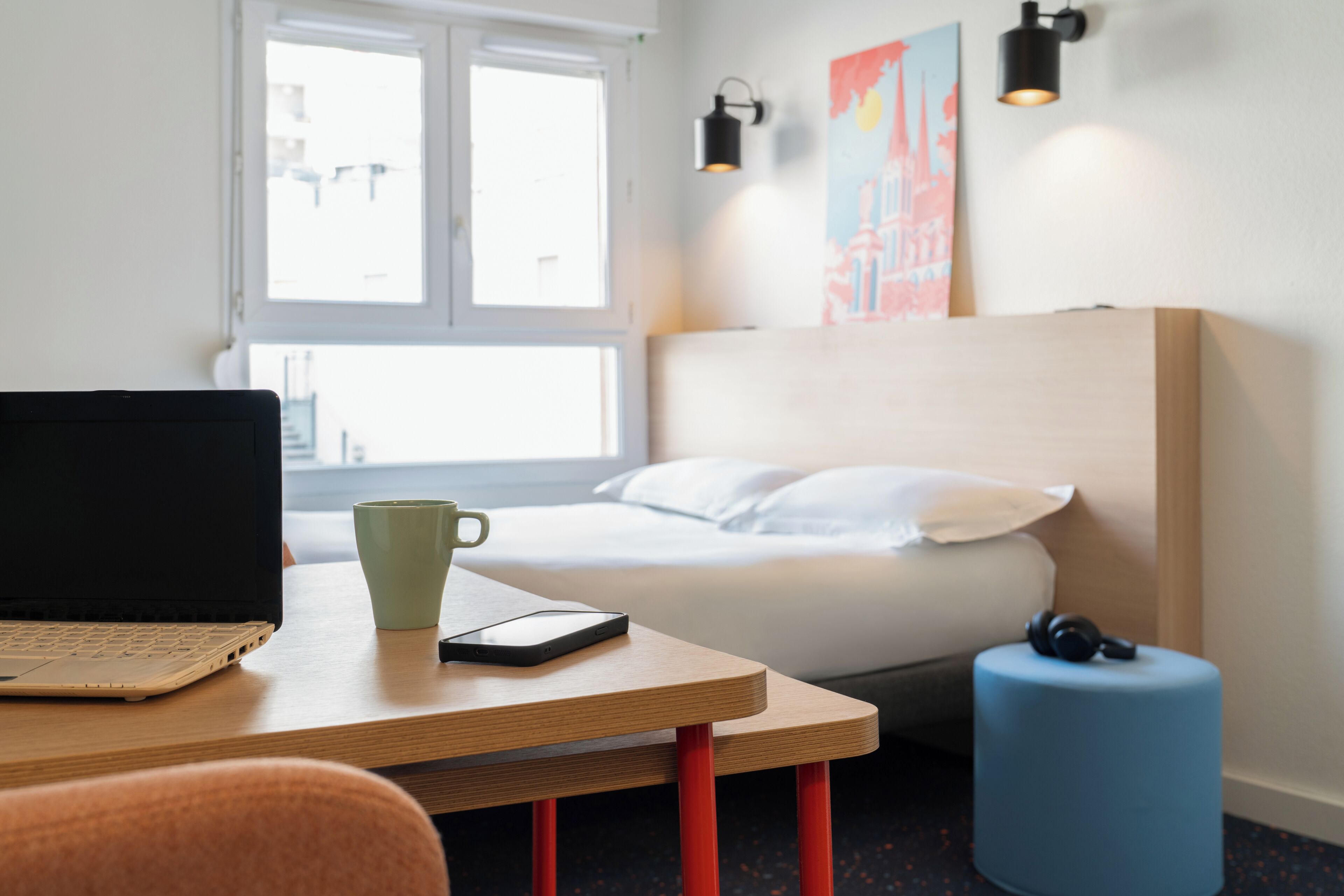 Una habitación de hotel con una cama, un escritorio con una computadora portátil, una taza, un teléfono inteligente, un reposapiés azul y una lámpara de pared.