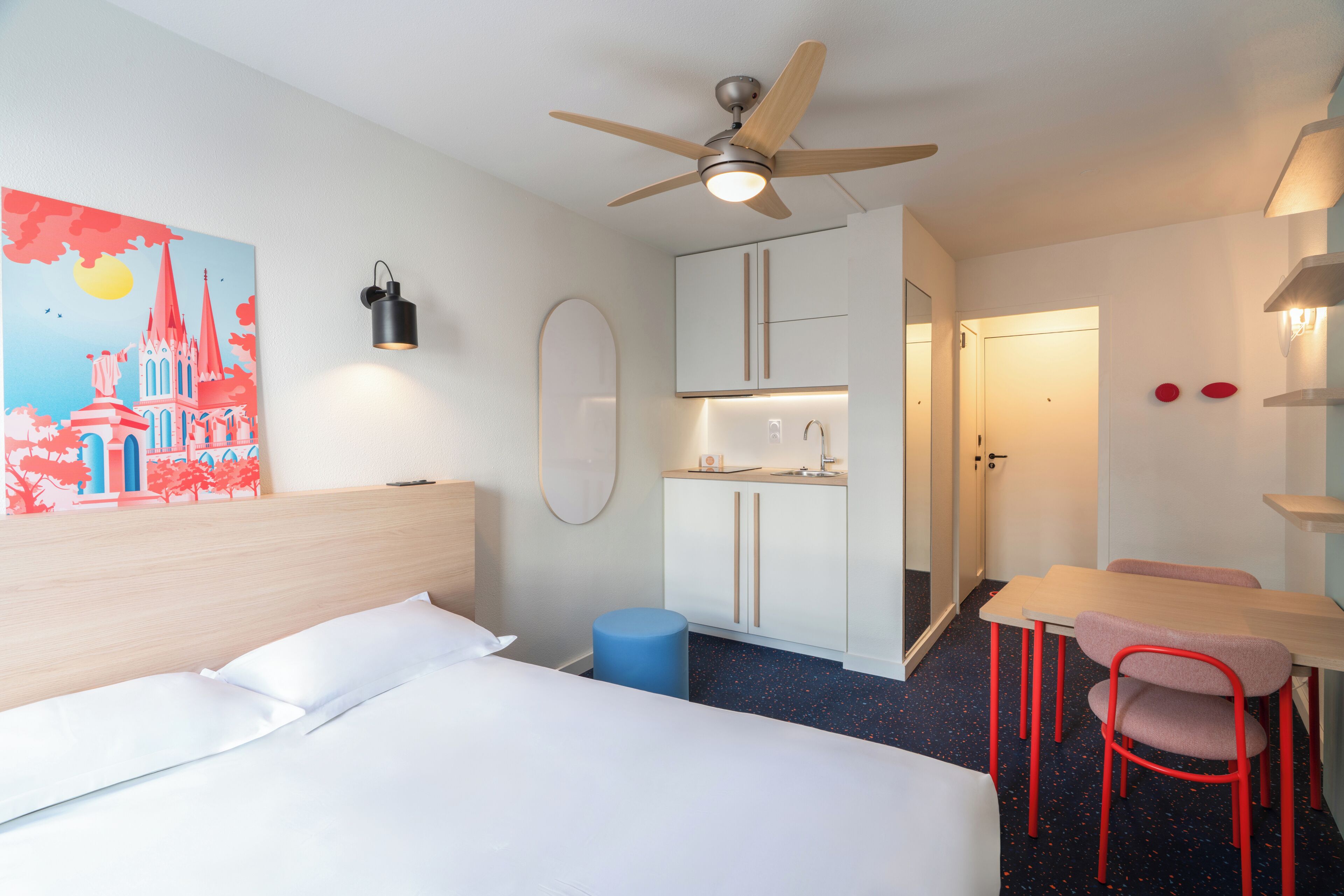 Een hotelkamer met een bed, een plafondventilator, een kitchenette en een eethoek met stoelen.