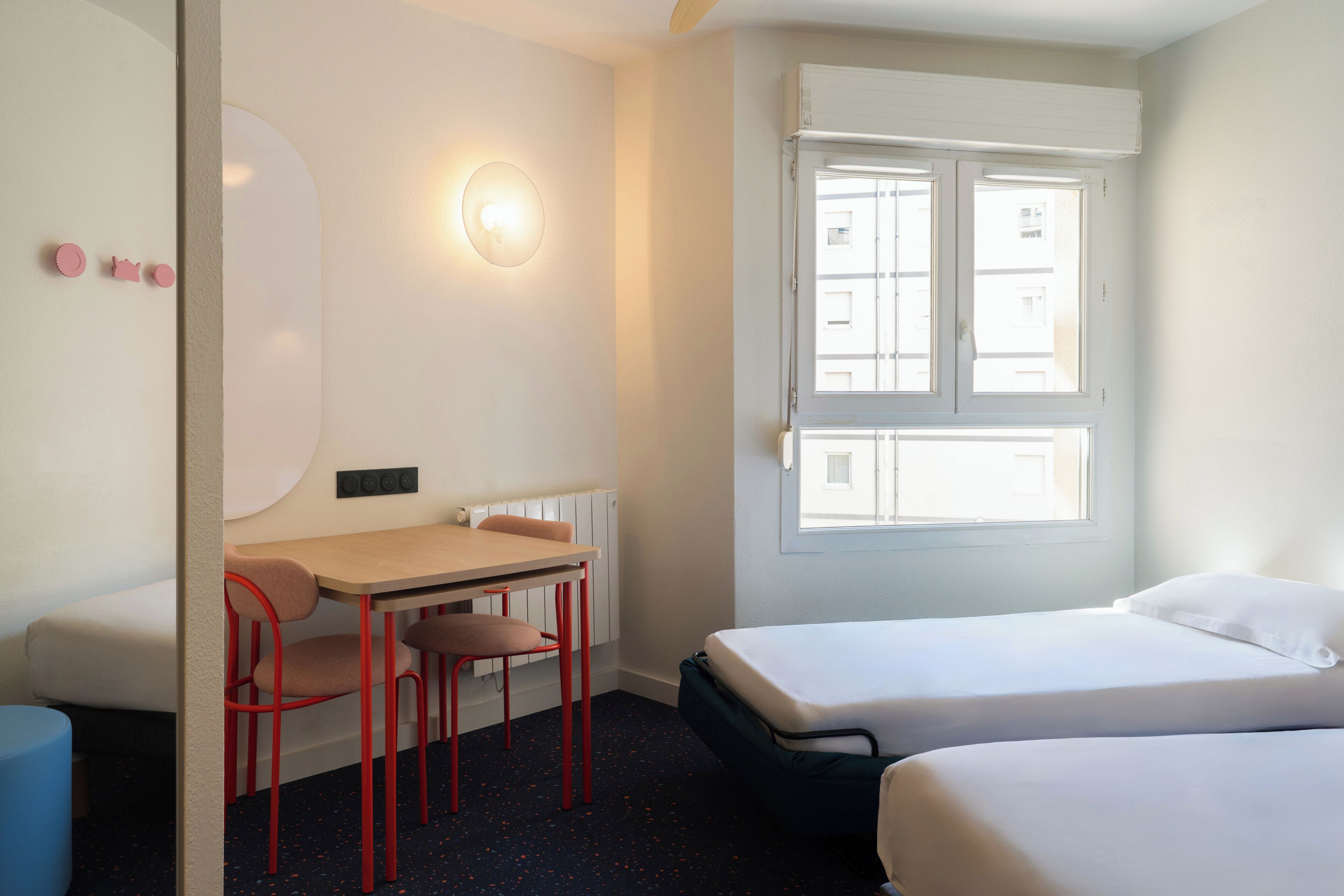 Une chambre avec deux lits, un bureau avec des chaises, une lampe fixée au mur et une fenêtre.