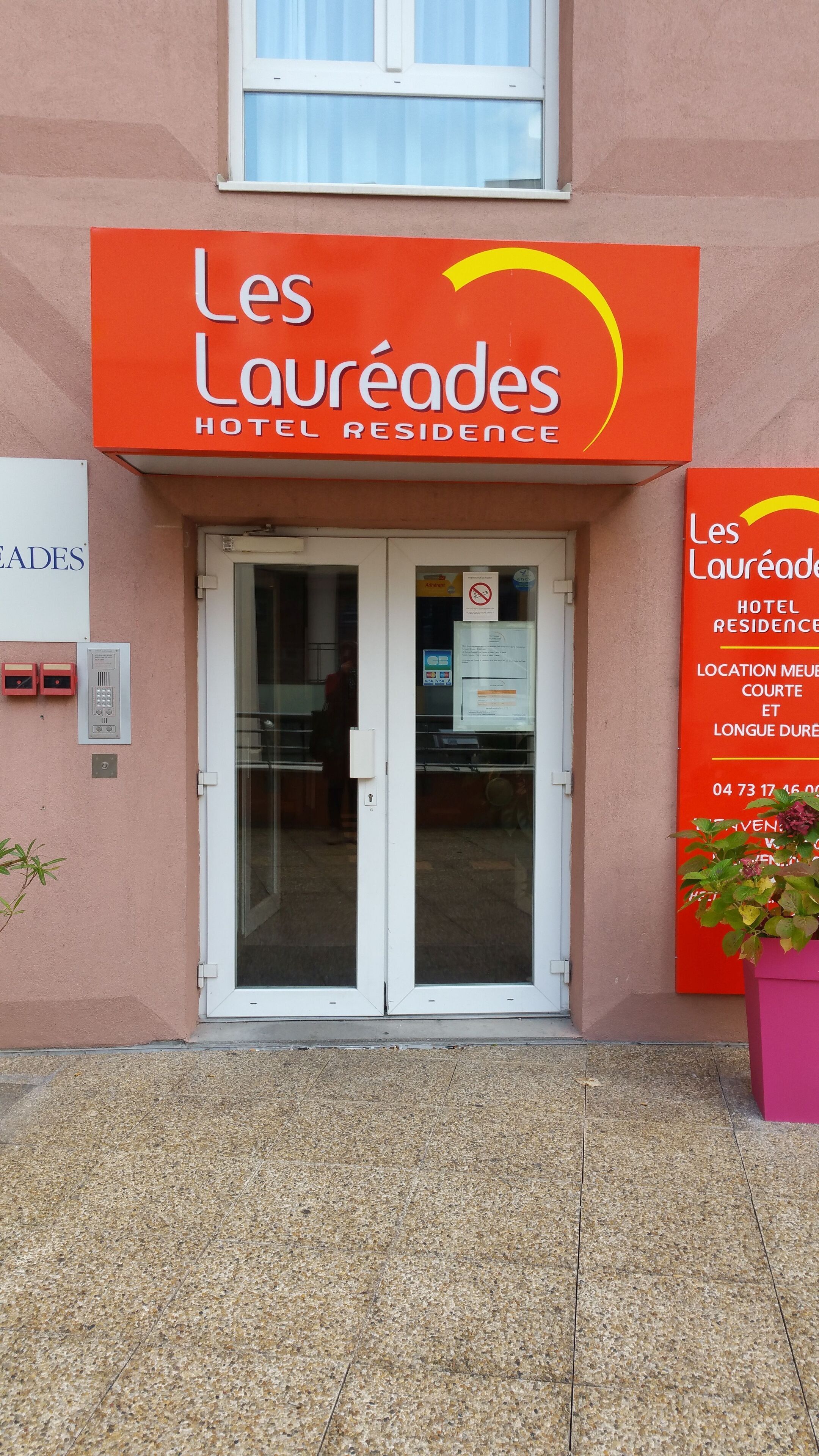 The exterior of Les Laureades Hotel Residence with a red sign, glass doors, and a small potted plant.