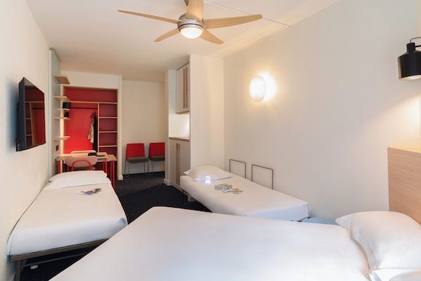 Habitación de hotel con tres camas individuales, ventilador de techo, televisor, escritorio y una silla roja.