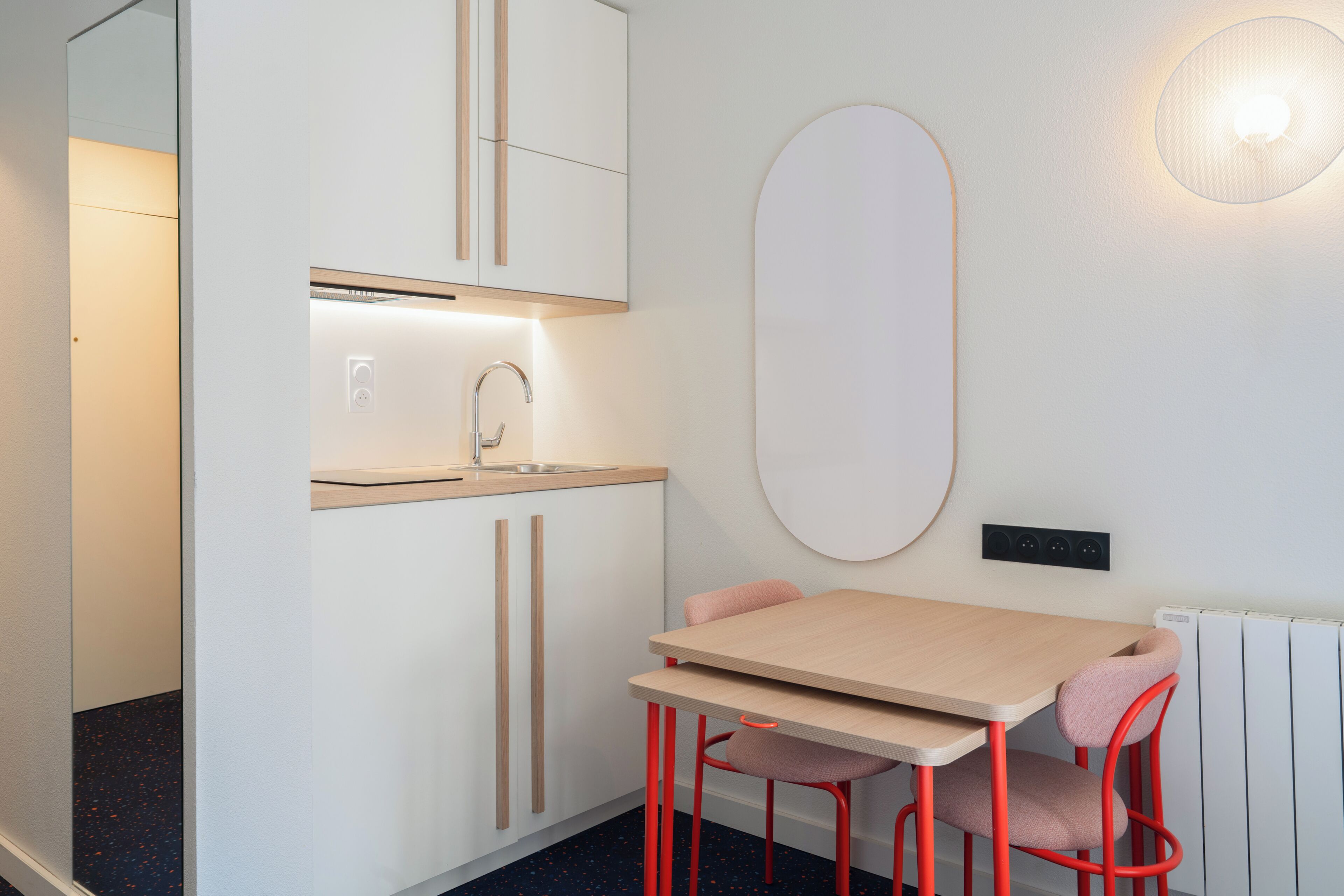 A modern kitchenette with a sink, cabinets, and a mirror. There is a table with two chairs and a radiator on the wall.