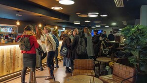 Un grupo de personas en una zona de bar con suelo de madera y una barra. Se ven luces en el techo y una pantalla de televisión al fondo.