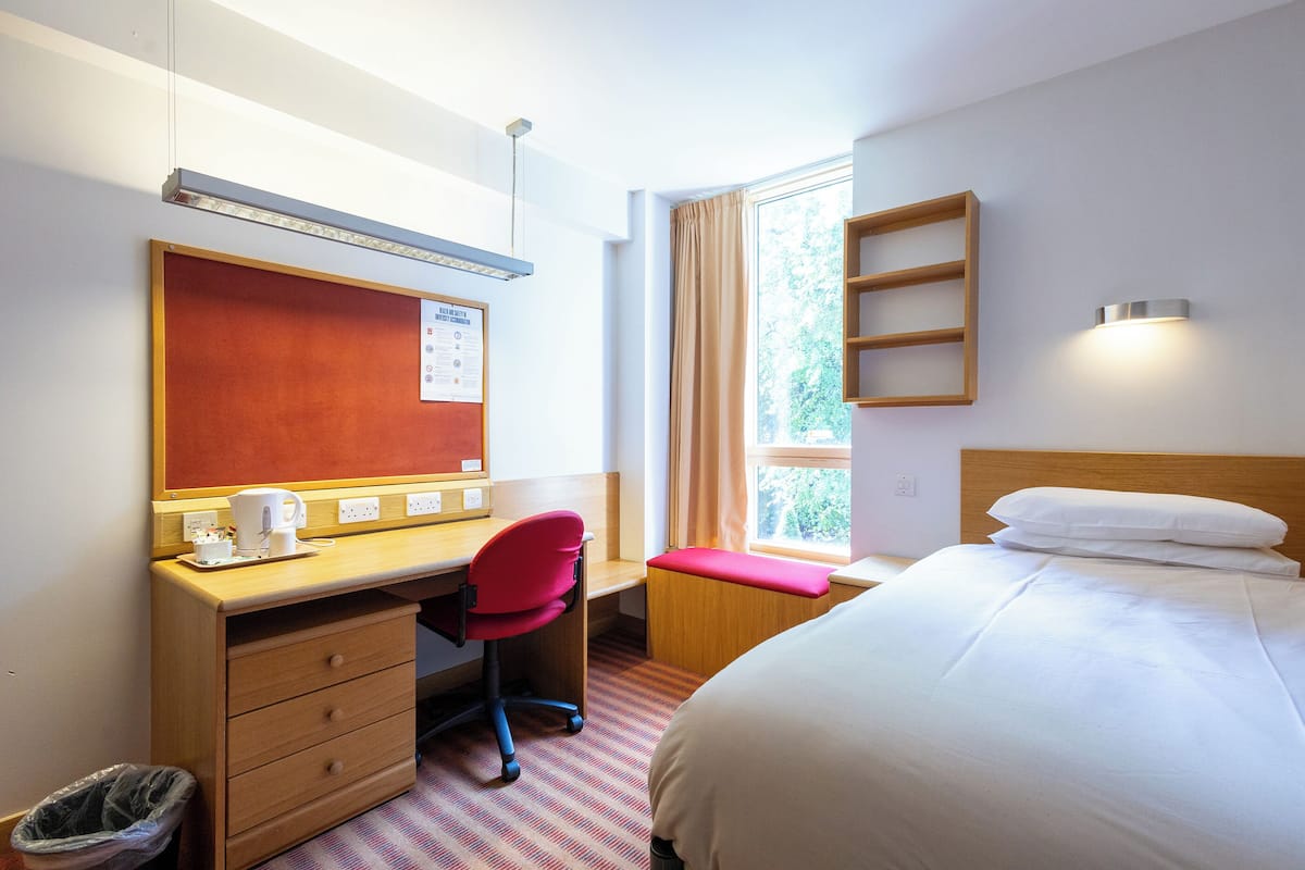 Quarto de hotel com cama, escrivaninha com cadeira, janela com cortinas, prateleira na parede e lixeira.