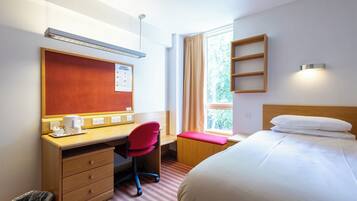 Een hotelkamer met een bed, een bureau met stoel, een raam met gordijnen, een wandplank en een prullenbak.