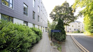 Un edificio moderno con un cartel que dice 'John Burnett House', un sendero pavimentado y abundante vegetación.