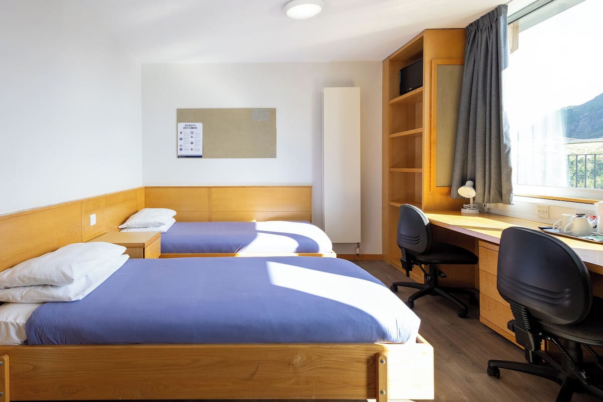 Quarto de hotel com duas camas, uma escrivaninha, uma cadeira e uma janela com cortinas.