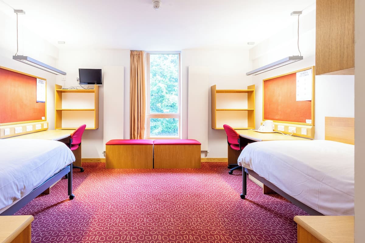 Quarto de hotel com duas camas, uma escrivaninha, uma cadeira, uma televisão e uma janela com cortinas.